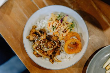 Lezzetli Teriyaki usulü tavuklu pilav kasesi taze salata ve ahşap bir masada gevrek soğan halkası. Yemek blogları için mükemmel.