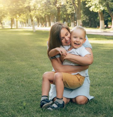 Mutlu genç anne, güneşli bir yaz gününde çimenlikteki parkta oğluna sarılır. Yakın plan. Duygu neşedir. Mutlu aile kavramı