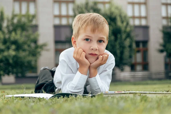 A pensive Caucasian boy lies on the grass with a book. Primary school student lies on the grass against the background of the school building. Hobbies and leisure. Reading in nature.
