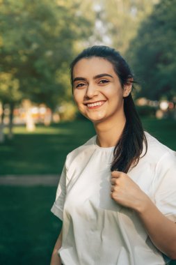 Beyaz tişörtlü, uzun siyah saçlı, güzel beyaz tenli bir esmer, güneşli bir yaz gününde yeşil ağaçların arkasında geniş bir gülümseme sergiliyor. Güzel gülüşlü, kaygısız bir Arap kadın.