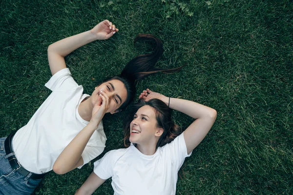 两个身穿白色T恤的年轻美丽的白人黑发姑娘躺在绿草上笑着 无忧无虑的女人夏天在草坪上聊天很开心 — 图库照片