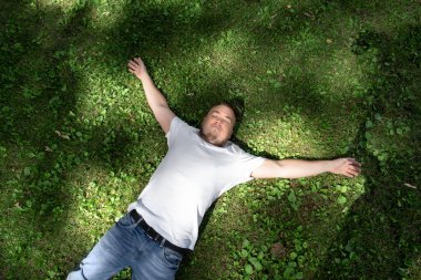 Kolları açık ve gözleri çimlere kapalı uzanmış yatarken gülümseyen yakışıklı, beyaz bir adam manzarası.