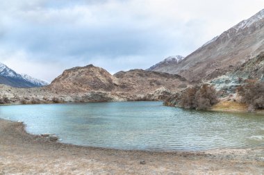 Yarab Tso 'nun manzarası, Hindistan' ın Nubra Vadisi 'ndeki turkuaz mavi göl. Durgun su, arka planda görkemli Himalaya dağlarıyla bulutlu gökyüzünü yansıtırken renkli Budist dua bayrakları ruhani ve kültürel bir dokunuş ekliyor..