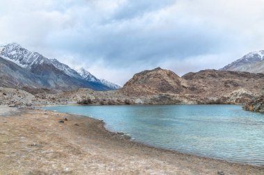 Yarab Tso 'nun manzarası, Hindistan' ın Nubra Vadisi 'ndeki turkuaz mavi göl. Durgun su, arka planda görkemli Himalaya dağlarıyla bulutlu gökyüzünü yansıtırken renkli Budist dua bayrakları ruhani ve kültürel bir dokunuş ekliyor..