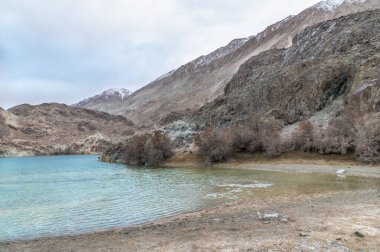 Yarab Tso 'nun manzarası, Hindistan' ın Nubra Vadisi 'ndeki turkuaz mavi göl. Durgun su, arka planda görkemli Himalaya dağlarıyla bulutlu gökyüzünü yansıtırken renkli Budist dua bayrakları ruhani ve kültürel bir dokunuş ekliyor..