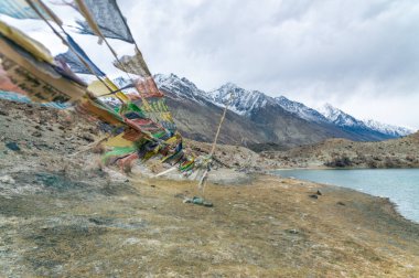 Yarab Tso 'nun manzarası, Hindistan' ın Nubra Vadisi 'ndeki turkuaz mavi göl. Durgun su, arka planda görkemli Himalaya dağlarıyla bulutlu gökyüzünü yansıtırken renkli Budist dua bayrakları ruhani ve kültürel bir dokunuş ekliyor..