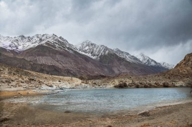 Yarab Tso 'nun manzarası, Hindistan' ın Nubra Vadisi 'ndeki turkuaz mavi göl. Durgun su, arka planda görkemli Himalaya dağlarıyla bulutlu gökyüzünü yansıtırken renkli Budist dua bayrakları ruhani ve kültürel bir dokunuş ekliyor.. 