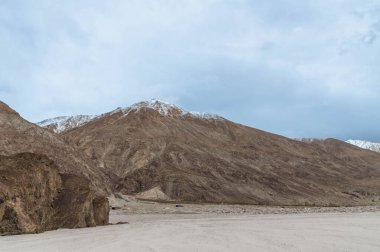 Yarab Tso 'nun manzarası, Hindistan' ın Nubra Vadisi 'ndeki yüksek rakımlı kutsal göl. Ön planda göl kıyısı boyunca beyaz kumlar, görkemli Himalaya dağları ise bulutlu bir gökyüzünün altında yükseliyor..