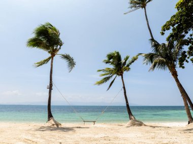 Palmiye ağaçları, turkuaz deniz ve mavi gökyüzüne bakan el değmemiş kumlu bir plajın üzerinde basit bir ahşap salıncak çerçevesi oluştururlar. Haad Yuan Sahili. Ko Pha Ngan, Tayland.