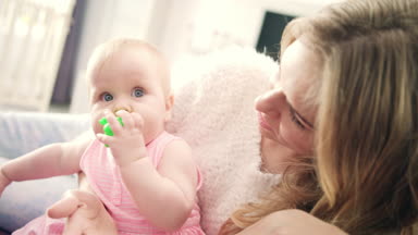 Anne kucağında oturan güzel bir kız bebek. Ağzında oyuncakla küçük bir kız.