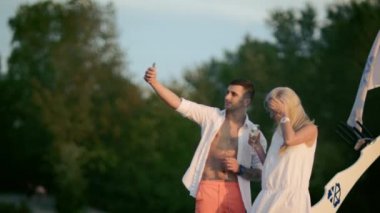 Güzel aşık selfie smartphone ile alıyor. Gün batımında aşk çift yapım resmi. Birlikte mutlu anları yakalamak. Holding bardak şampanya ve kameraya bakıyor gençler