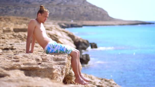 Snygg kille sitter på steniga stranden nära havet. Drömmer man tittar på havet — Stockvideo