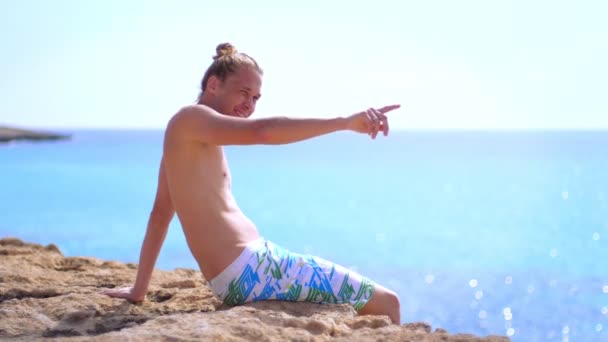 Турист показывает палец на берегу моря. Голый мужчина с грудью загорает — стоковое видео