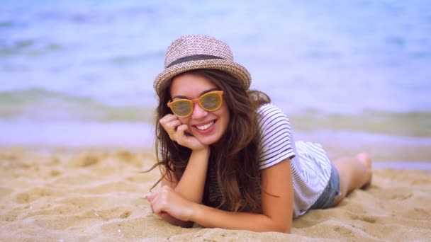 Chica con estilo en gafas de sol y sombrero moderno que pone en la arena disfrutando de vacaciones — Vídeos de Stock