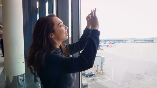 Modern girl taking film on phone departure of plane at airport throught window — Stock Video