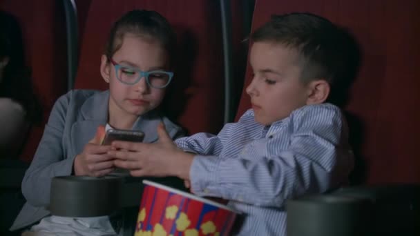 Niños comiendo palomitas de maíz en el cine. Niños usando teléfono móvil en el cine — Vídeo de stock