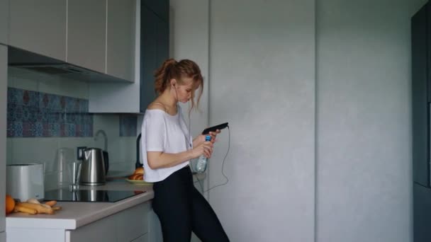 Passar flicka i sportkläder dricksvatten i köket. Kvinnan i vit t-shirt — Stockvideo