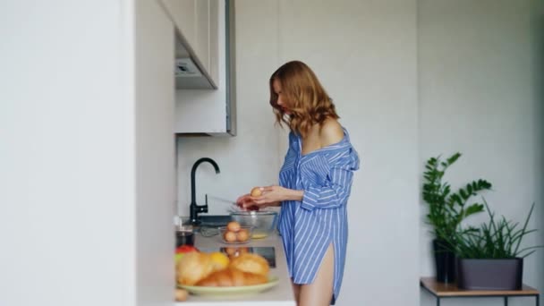 Menina sexy na camisa quebrando ovos em tigela de vidro na cozinha. Mulher muito jovem — Vídeo de Stock