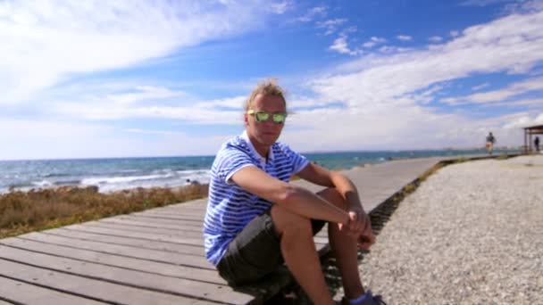 Un tipo moderno con gafas de sol sonriendo en la orilla del mar. Hombre con estilo en la costa del mar — Vídeo de stock