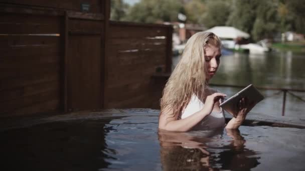 屋外のジャグジーでタブレットを使用してブロンドの女の子。水でタブレットを使用して金髪の女性 — ストック動画