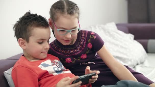 Юніорки хлопчик і дівчинка дивляться мобільний телефон вдома. Концепція щасливого дитинства — стокове відео