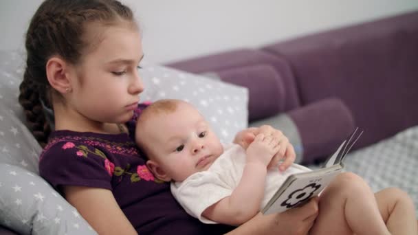 Menina bonita lendo livro infantil com bebê em casa. Irmã segurando irmão bebê — Vídeo de Stock