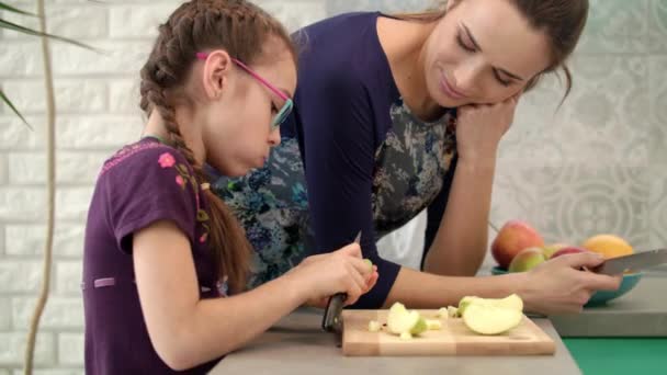 Chica comiendo manzana con madre en la cocina. Chica cortando manzana con cuchillo — Vídeos de Stock