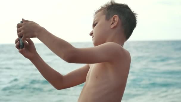 Joven niño filmando vídeo en el teléfono móvil en la playa del mar. Niño haciendo video — Vídeos de Stock