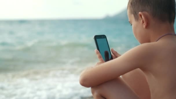 Маленький мальчик пользуется мобильным телефоном на берегу моря. Парень смотрит на мобильный телефон на пляже — стоковое видео