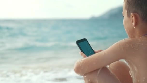 Splashes from sea waves fall on boy with phone in hands. Relaxing on resort — Stock Video