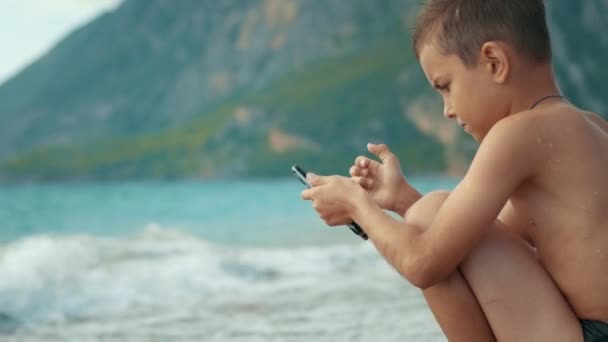 Nadenkende jongen met smartphone in de buurt van zee golven. Kind SMS berichten op telefoon — Stockvideo