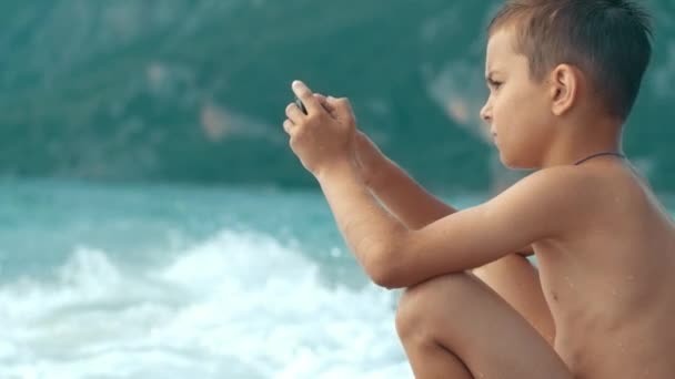 Niño jugando juegos en el teléfono inteligente mientras está sentado en la playa. Adolescente usando smartphone — Vídeo de stock