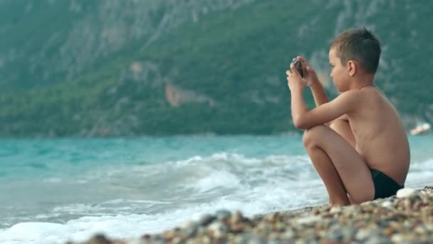 Ребенок наслаждается отдыхом в море. Мальчик играет в видеоигры на мобильном телефоне на пляже — стоковое видео