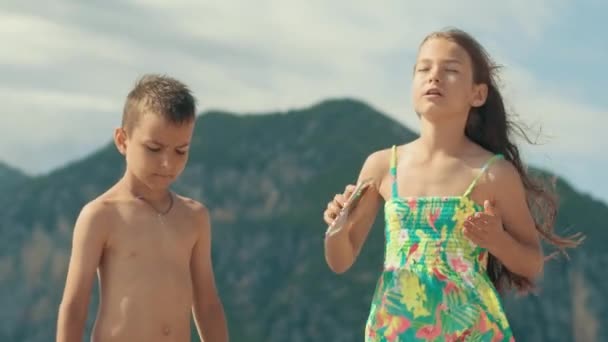 Dos niños se divierten escuchando música en el teléfono inteligente. Niño y niña bailando en la playa — Vídeos de Stock