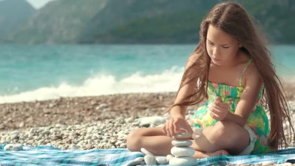 Chica concentrada construyendo torre de piedras en la playa del mar. Niño reflexivo soñando — Vídeo de stock