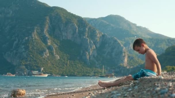 Niño sentado en la playa y lanzando arena y guijarros al mar — Vídeos de Stock