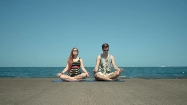 Νεαρό ζευγάρι που διαλογίζεται και κάθεται στην προβλήτα δίπλα στην παραλία. Διακοπές με μαθήματα γιόγκα — Αρχείο Βίντεο