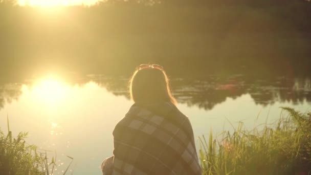 Chica solitaria en cuadros sentada en la orilla del río al atardecer. Mujer solitaria — Vídeos de Stock