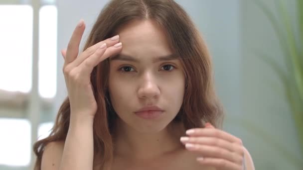Junge Frau sieht Gesicht im Spiegel im Badezimmer. Frau quetscht Akne — Stockvideo