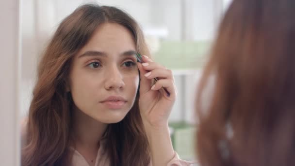 Menina atraente arrancando sobrancelhas no rosto com pinças espelho frontal no banheiro — Vídeo de Stock