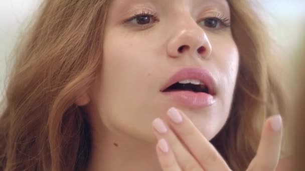 Красивая женщина наносит крем на кожу лица во время макияжа перед зеркалом в ванной комнате — стоковое видео