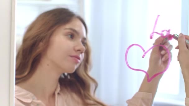 Mujer joven escribiendo por lápiz labial palabra amor en el espejo con el corazón en el baño — Vídeos de Stock