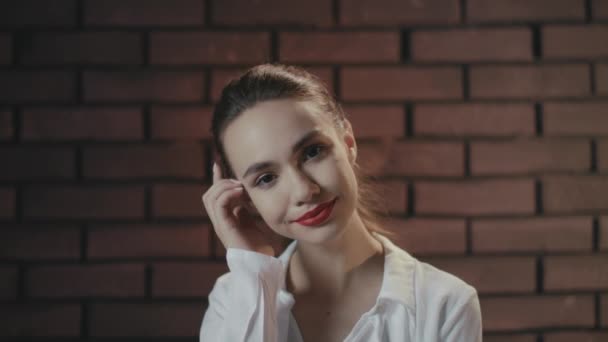 Стильна дівчина торкається волосся і вуха, позуючи в студії з цегляною стіною — стокове відео
