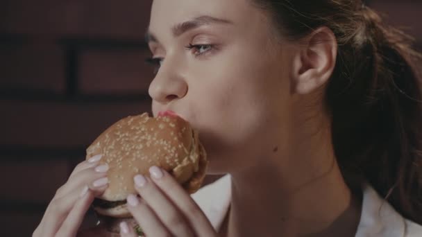 Hladová žena jíst burger na cihlové zdi pozadí v fast food kavárně — Stock video
