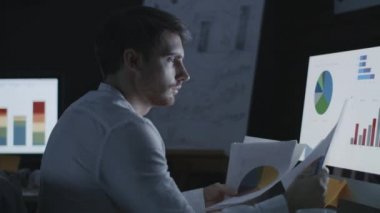 Karanlık ofisteki finansal çizelge ve grafiklere bakan bir iş adamı.