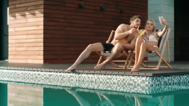 Радісна пара фотографує селфі біля басейну. Веселі люди насолоджуються коктейлем — стокове відео