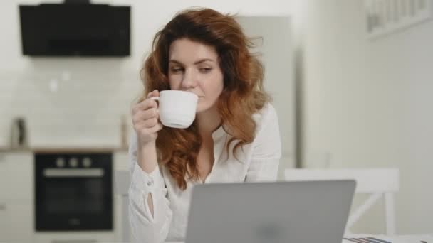 Heureuse femme travaillant ordinateur portable sur un lieu de travail éloigné. Jeune femme boire du thé à la maison . — Video