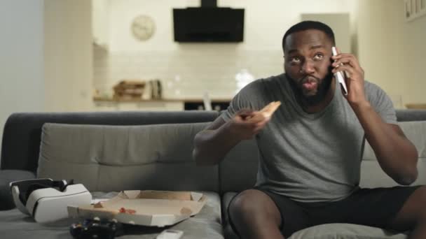 Hombre negro comiendo pizza delante de la televisión. Varón fan comentando resultados del partido — Vídeo de stock