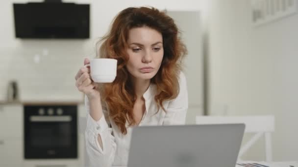 Концентрированная женщина пьет чай на утренней кухне. Молодая леди работает ноутбук — стоковое видео