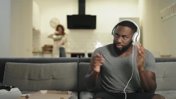 Усміхнений чорний чоловік танцює в навушниках на сучасній кухні . — стокове відео